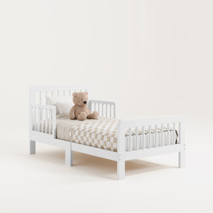Angled view of a white toddler bed with bedding and teddy bear